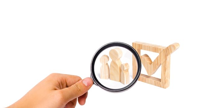 Magnifying glass is looking at the Three wooden human figures stand together next to a tick in the box. The concept of elections and social technologies. Volunteers, parties, candidates. Human rights