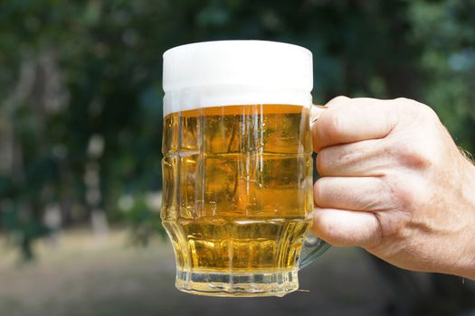 glass mug with beer and foam in a man's hand on the background of nature close up