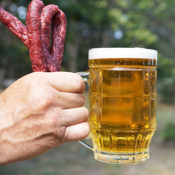 glass mug with beer and sausages in a man's hand on the background of nature close up