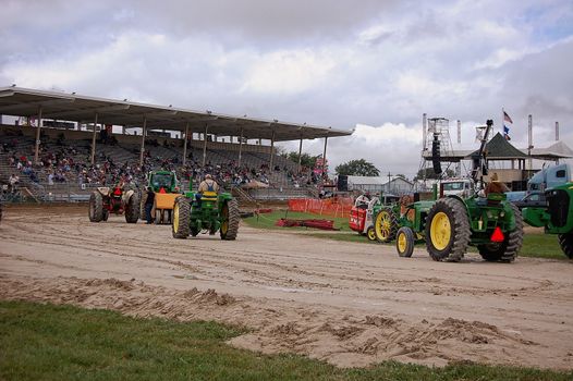 This is a group of tractor's awaiting there turn during a tractor pull at a county fair.