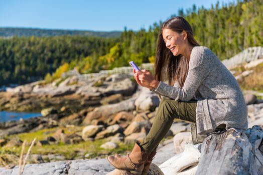 Happy girl texting sms message on mobile phone on nature forest hike in autumn outdoors. Asian woman playing on cellphone.