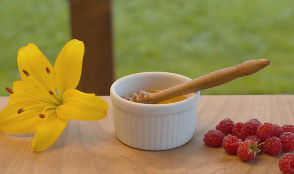 Close up honey spoon in a bowl with honey. Raspberry and lily on a table. Healthy fresh food concept. Summer still life.