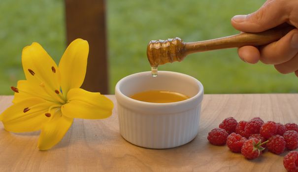 Close up hand of a man taking honey with honey spoon. Honey pouring into a bowl. Raspberry and lily on a table in the garden at summer day. Healthy fresh food concept.