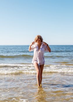 young blonde woman in a wet white shirt coming out of the water near the seashore on sunny summer day