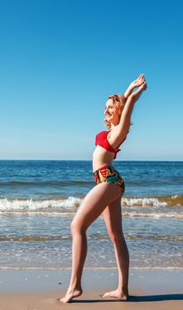 young blonde girl in a red swimsuit standing on the seashore with her hands behind her head on sunny summer day