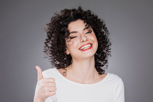Happy smiling excited woman giving thumb up. Caucasian female with curly hair anf toothy smile on gray background