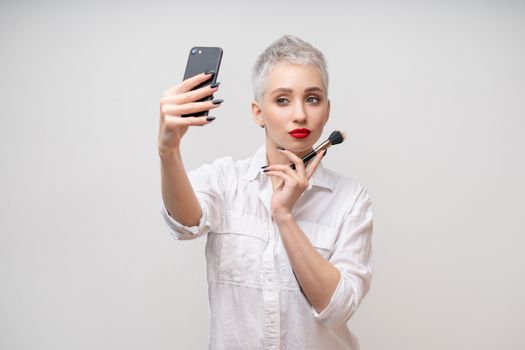 Studio portrait of beautiful trendy girl with short hair taking selfie via cell phone over white isolated background Vloger Blogger Makeup artist
