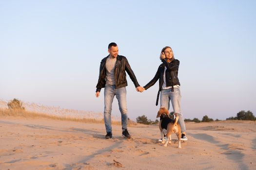 Young beautiful caucasian couple wearing leather jacket and jeans walks desert sand with Beagle dog best friend. Family with dog without children resting in nature. positive emotions happy people