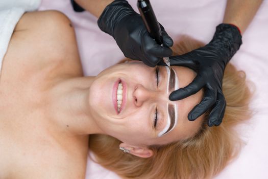 Cosmetology cabinet client lies on couch. Beautician applies permanent ink on eyebrows. Procedure of permanent eyebrow makeup. Close up black glove. Beauty industry