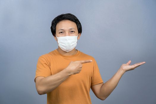 Asian man wearing face surgical mask pointing and presenting some product about coronavirus on blue color background, covid19 outbreak and pandemic,healthcare and protection against virus concept