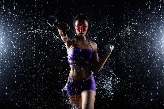 Beautiful young girl in purple sportswear poses with dumbbells in aqua studio. Drops of water spread about her fitness body. The perfect figure on the background of water splashes.