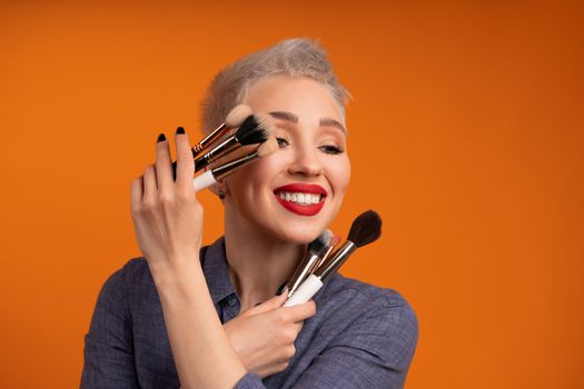 Close up portrait makeup artist. Make up courses. Concept of self visage masterclasess. Woman hold makeup brushes on the hands. Studio shot on the orange color background. Positive people smiling