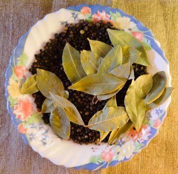 top view of a composition of black, white and red hot pepper and bay leaves on a plate with painted flowers, background - rough fabric