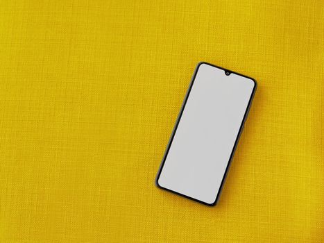 Black mobile smartphone mockup lies on the surface with a blank screen isolated on a yellow fabric background. Top view close up with selective focus and copy space.