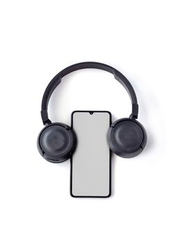 Black wireless headphone and mobile smartphone with a blank screen mockup lay on the surface of a white background. Top view flat lay with copy space. Music concept. The headphones look like ears.