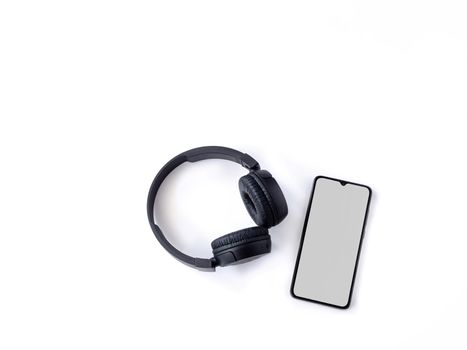 Black wireless headphone and mobile smartphone with a blank screen mockup lay on the surface of a white background. Top view flat lay with copy space. Music concept.