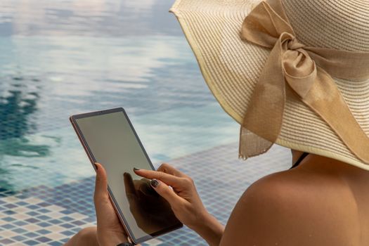 Vacation lifestyle scene of young woman in black bikini using tablet while sitting by the pool in morning time. Weekend and holiday lifestyle, Relaxing on the poolside.