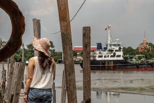 The back of a young woman in casual clothes standing on an old wooden pier and looking at the view Chao Phraya River the afternoon.  Personal holiday.