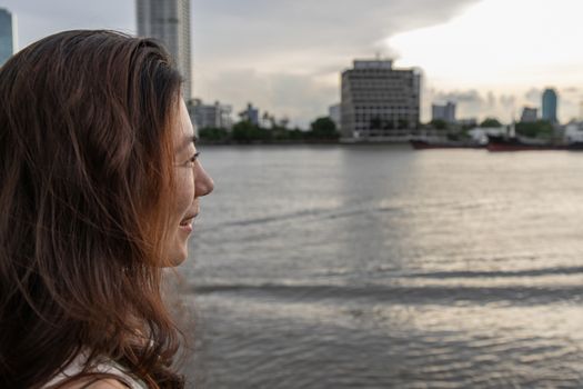 Side view of young woman standing by river and smiles happily in the scenery on the chao phraya river in the evening.