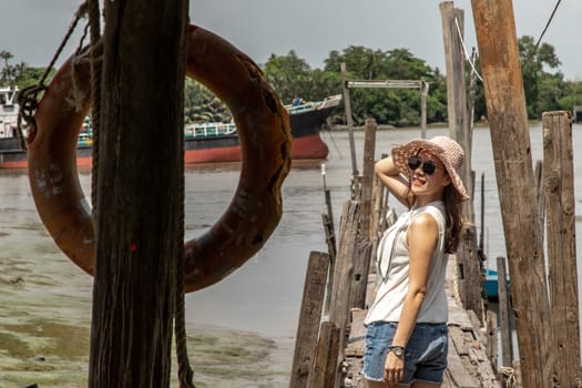 The back of a young woman in casual clothes standing on an old wooden pier and looking at the view Chao Phraya River the afternoon.  Personal holiday.