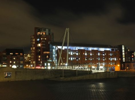 a cityscape view of the canal entrance to the clarence dock area of leeds with a pedestrian bridge crossing the water with reflections of lights and buildings against a night sky with clouds