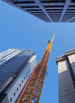 vertical view of a tall yellow construction crane working on a new concrete building with large surrounding towers