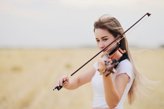 Beautiful romantic girl with loose hair playing the violin in the field after the harvest. Square sheaves of hay in the field. Violin training