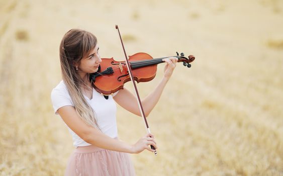 Beautiful romantic girl with loose hair playing the violin in the field after the harvest. Square sheaves of hay in the field. Violin training