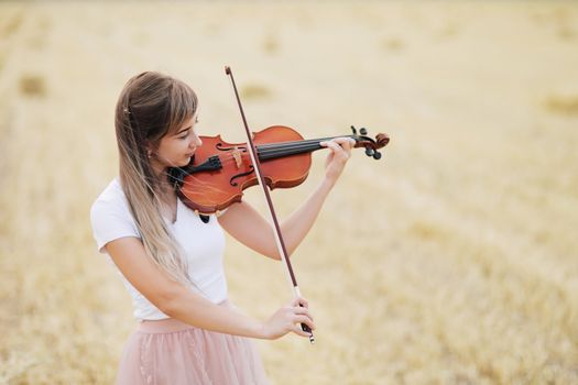 Beautiful romantic girl with loose hair playing the violin in the field after the harvest. Square sheaves of hay in the field. Violin training