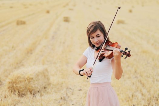 Beautiful romantic girl with loose hair playing the violin in the field after the harvest. Square sheaves of hay in the field. Violin training