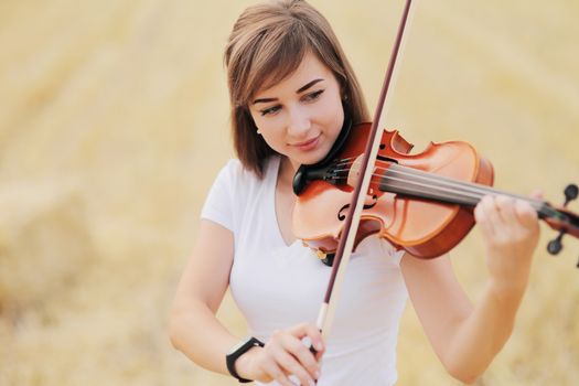 Beautiful romantic girl with loose hair playing the violin in the field after the harvest. Square sheaves of hay in the field. Violin training