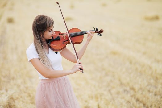 Beautiful romantic girl with loose hair playing the violin in the field after the harvest. Square sheaves of hay in the field. Violin training