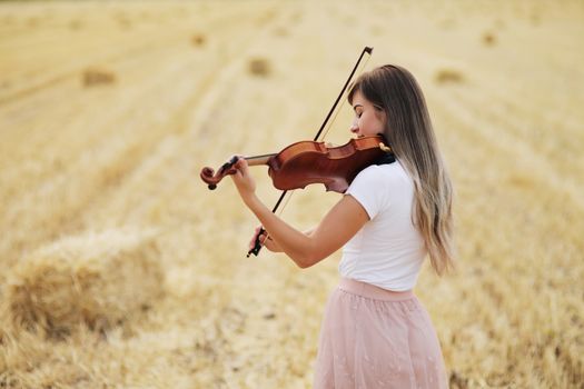 Beautiful romantic girl with loose hair playing the violin in the field after the harvest. Square sheaves of hay in the field. Violin training