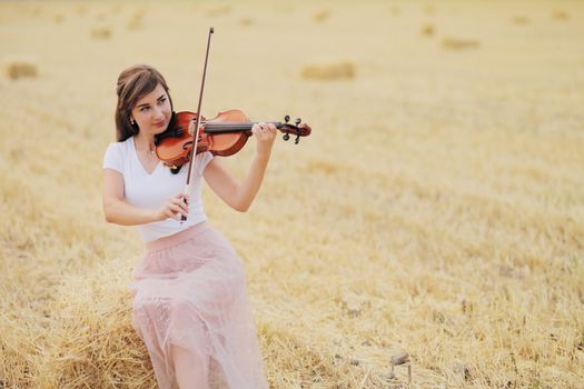 Beautiful romantic girl with loose hair playing the violin in the field after the harvest. Square sheaves of hay in the field. Violin training