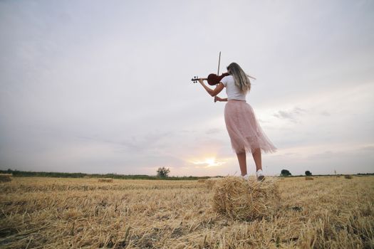 Beautiful romantic girl with loose hair playing the violin in the field after the harvest. Square sheaves of hay in the field. Violin training