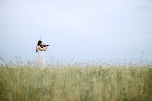 Beautiful romantic girl with loose hair playing the violin in the field after the harvest. Square sheaves of hay in the field. Violin training