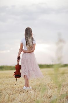 Romantic young woman with flowing hair, holding a violin in her hand in a field after harvest. Square sheaves of hay in the field. Violin training