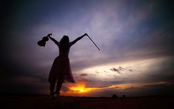 Romantic young woman with loose hair, holding a violin in her hand in a field at sunset after harvest. Square sheaves of hay in the field. Violin training