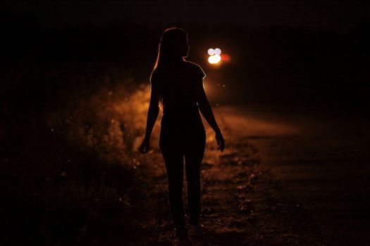 Silhouette of young slender woman in the backlight of car headlights on the auto road at night. The girl is hitchhiking.