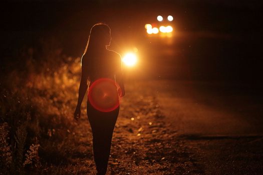 Silhouette of young slender woman in the backlight of car headlights on the auto road at night. The girl is hitchhiking.