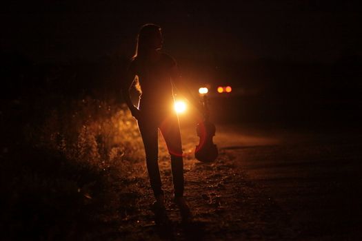 Silhouette of a slender young woman with violin in car headlights illumination. A young slender girl in the backlight of car headlights on the auto road at night. The girl is hitchhiking.