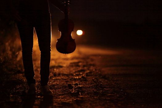 Silhouette of a slender girl's legs and a violin in the backlight of headlights. A young slender woman in the backlight of car headlights on the auto road at night. The girl is hitchhiking.