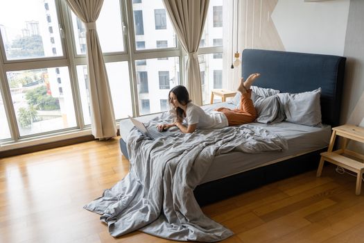 A beautiful young brunette girl is working on the laptop while lying on a bed by a panoramic window with a beautiful view from a high floor. Stylish modern interior. A cozy workplace.