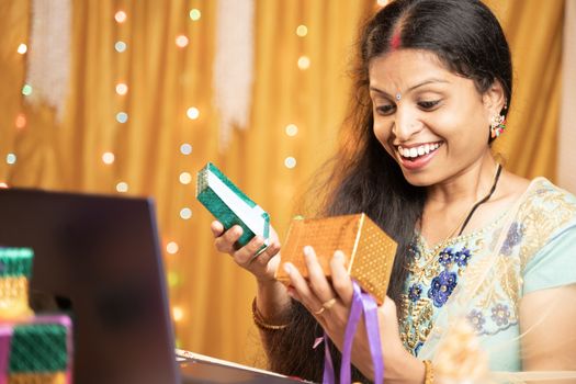 Happy excited Woman opening gift infront of laptop during video call or chat at Raksha Bandhan festival ceremony - Concept of distance relations, celebrations and lifestyle