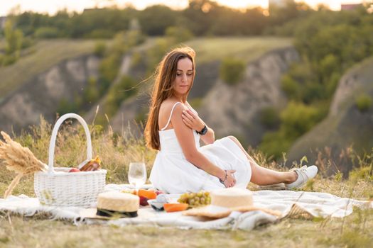 Gorgeous young brunette girl in a white sundress having a picnic in a picturesque place. Romantic picnic.