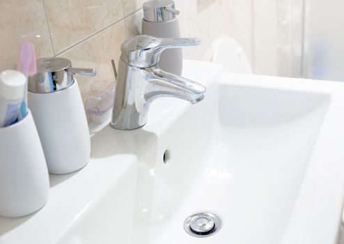 a white ceramic sink with a chromed tap with soap and toothbrush containers in a bathroom. Interior of a house.