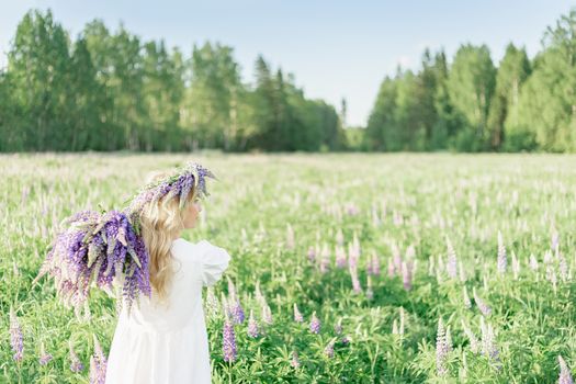 A girl with a bouquet of flowers over her shoulder in a white dress and a flower wreath walks back along the flower field, in the flower field of lupins. Lupine field and the girl backs away