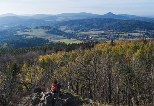 Young woman hiker takeing pictutre of Luzicke hory panorama from Hochwald Hvozd view-point of the Lusatian Mountains with autumn colored deciduous and coniferous tree forest and green hills, golden hour light.