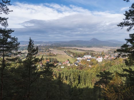 View from Hrabencina vyhlidka lookout on village Sloup v cechach in luzicke hory, Lusatian Mountains with autumn colored deciduous and coniferous tree forest and green hills, blue sky, white clouds.
