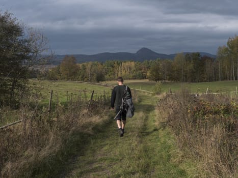 young man walking on dirt road at meadow with autumn colorful forest and trees and hills, moody sky. Landscape in luzicke hory Lusatian mountains